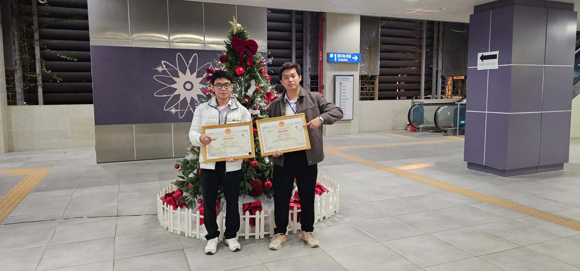 Hai người cầm bằng khen bên cây thông trang trí.