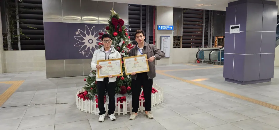 Two people holding award certificates beside a decorated Christmas tree.